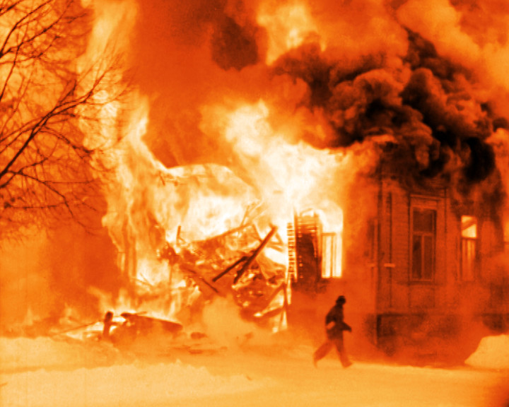 A wooden house burns with high flames and black smoke while a figure runs past through snow.
