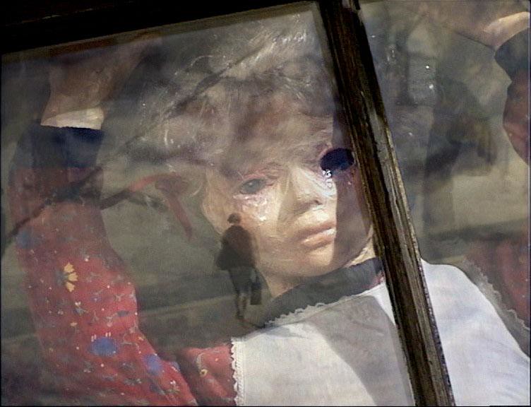 A puppet girl in a red flowered dress presses against a window, a walking figure reflected in the glass.