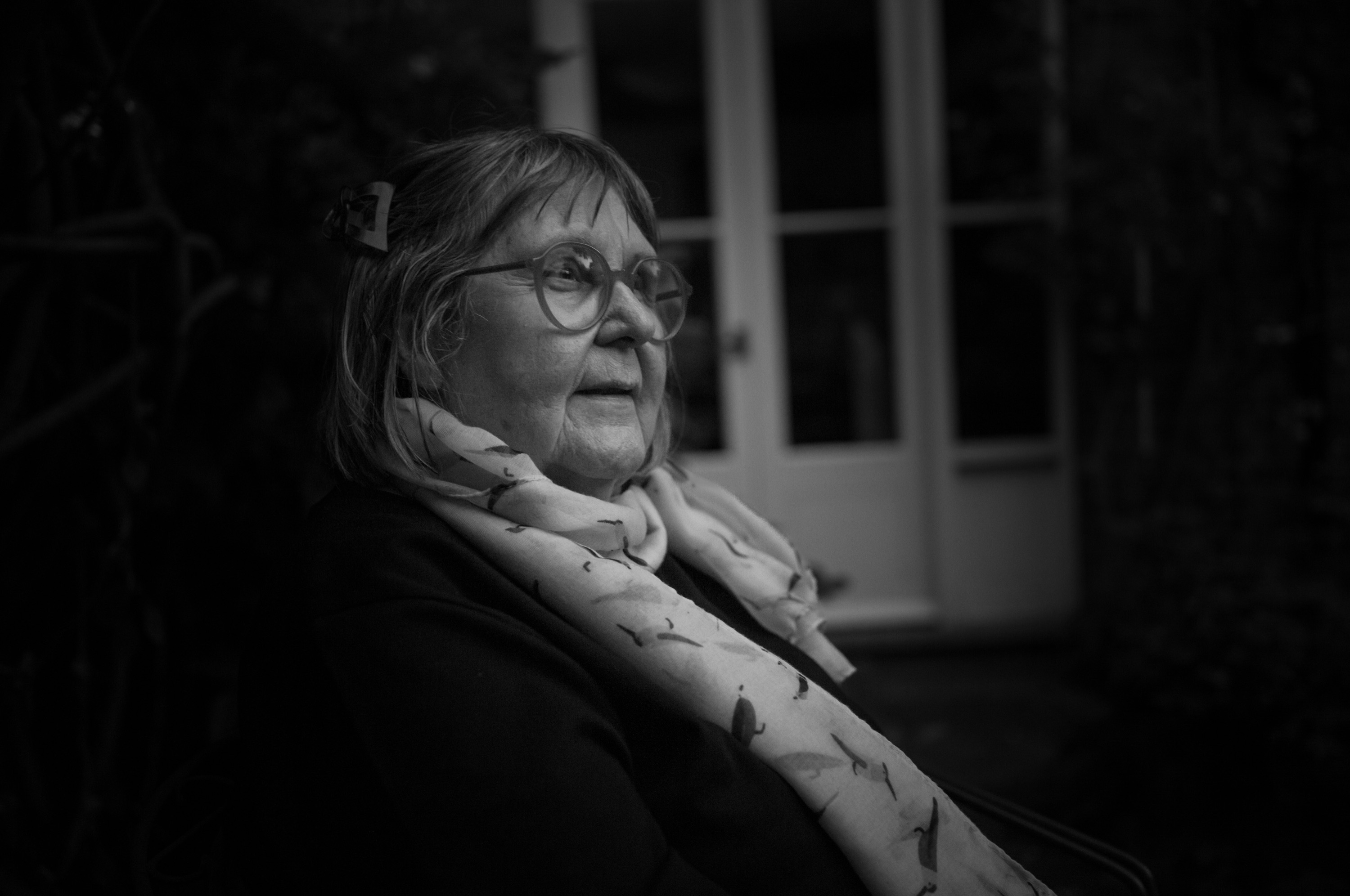Marjut Rimminen in round glasses and a patterned scarf, seated by the French doors at dusk