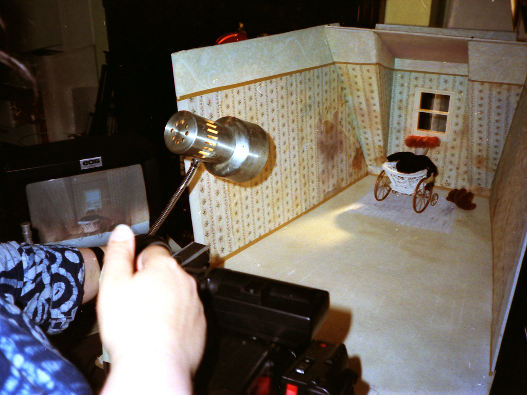 A hand aims a camera at a miniature wallpapered room with a pram and a brown stain.