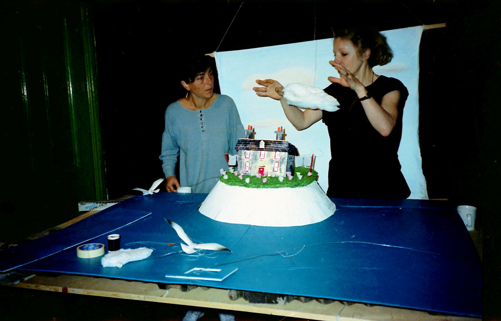 One woman holds a wisp of cotton wool cloud above a model house; another watches.