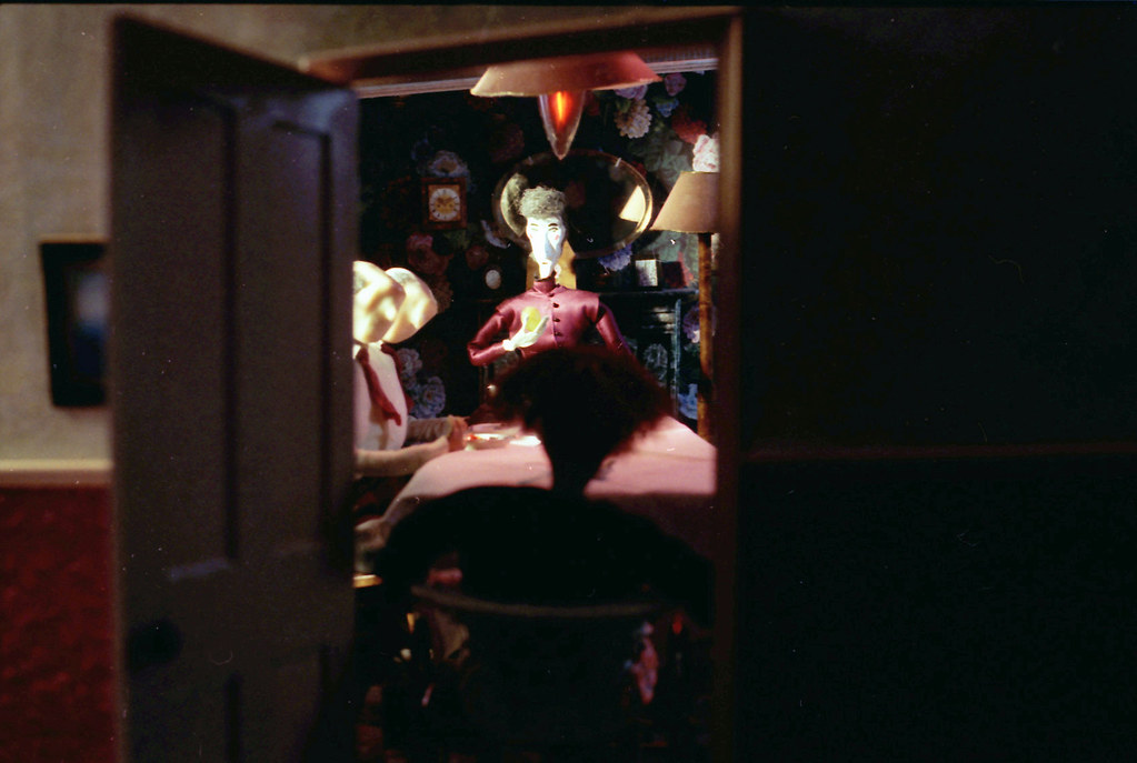 Through a half-open door, puppets sit at a dining table under a red hanging lamp.