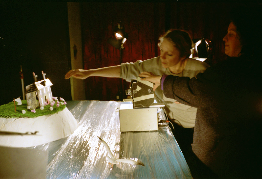 Two women work over a model island set in a sea of crumpled foil, under a lamp.
