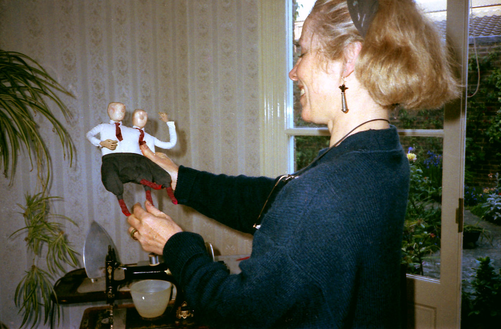 A woman holds up a puppet with two identical bald heads in white shirts and red ties.