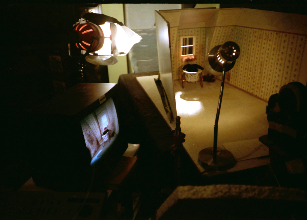 Studio lamps and a monitor surround a lit miniature room where a pram stands by the wall.