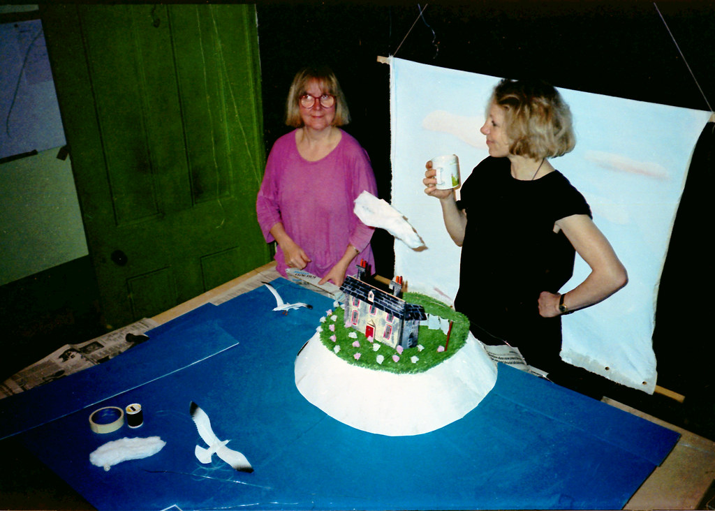 Two women stand behind a model house on a white cliff, blue paper sea and painted sky.