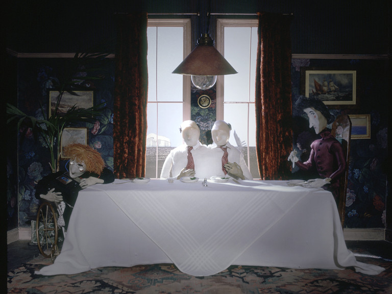 Still from The Stain, four puppet figures sitting around a white dining table under a hanging lamp, tall curtained windows behind them