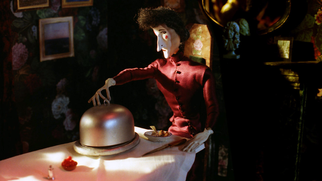 A puppet in a dark red dress reaches towards a domed silver cover on a laid table.
