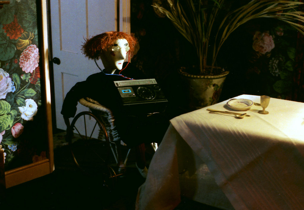 A red-haired puppet in round glasses sits in a wheelchair with a radio on its lap.