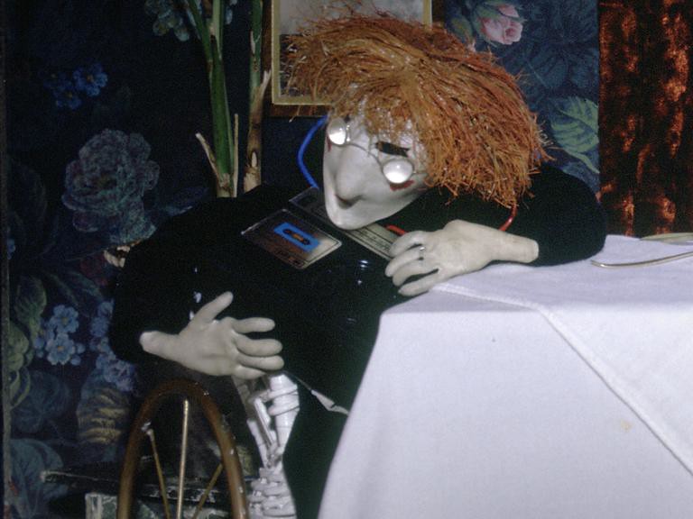 A red-haired puppet in round glasses leans on the tablecloth holding a personal cassette player and headphones.