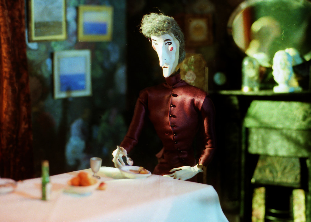 A puppet woman in a dark red high-collared dress eats alone at a white-clothed table.