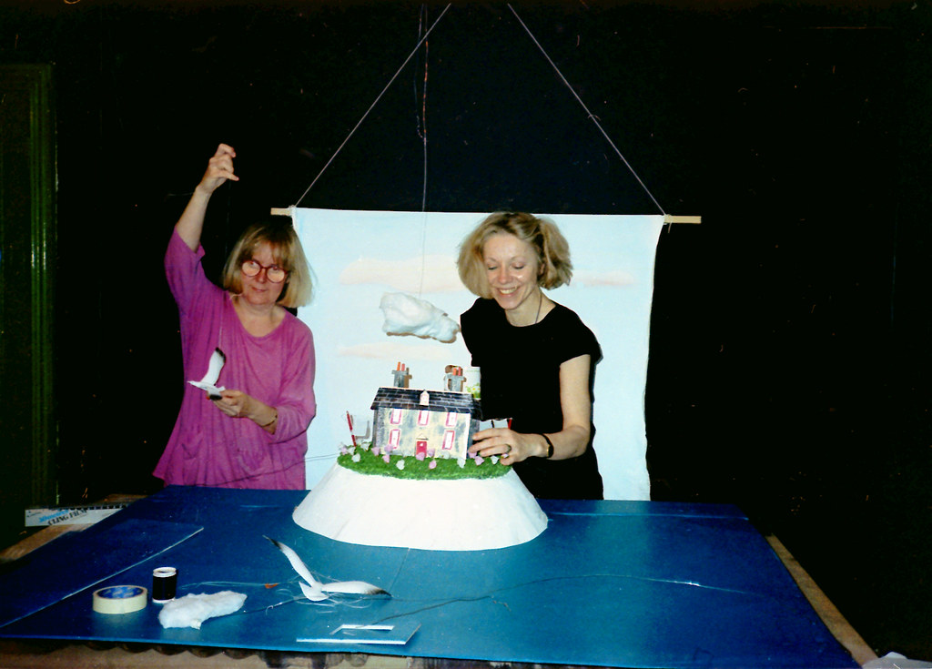 One woman raises a thread overhead while another steadies a model house on a white cliff.