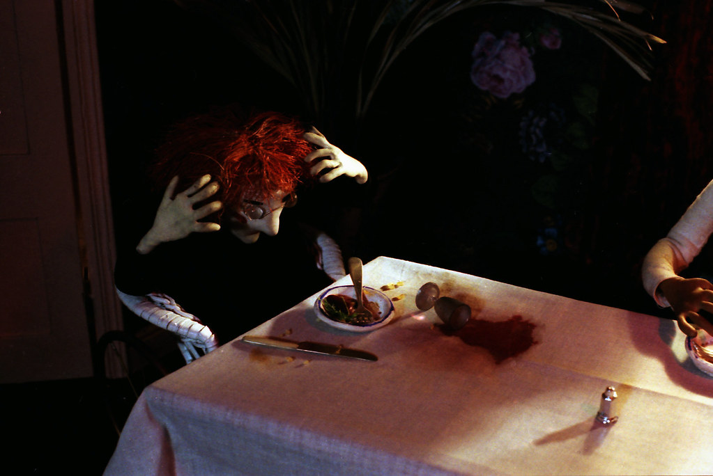 A red-haired puppet clutches its head beside a spreading red stain on the white tablecloth.
