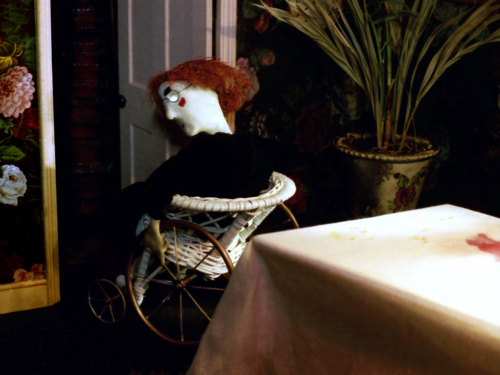 A red-haired puppet in black sits in a wicker chair on wheels beside a doorway.