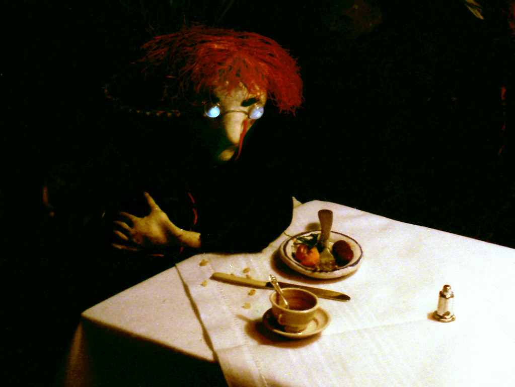 A red-haired puppet, its glasses catching the light, leans over a table set with food.