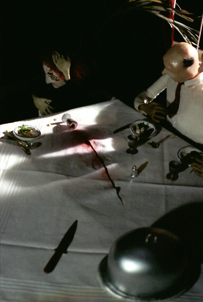 A red stain, a fallen goblet and a knife on the tablecloth between two seated figures.