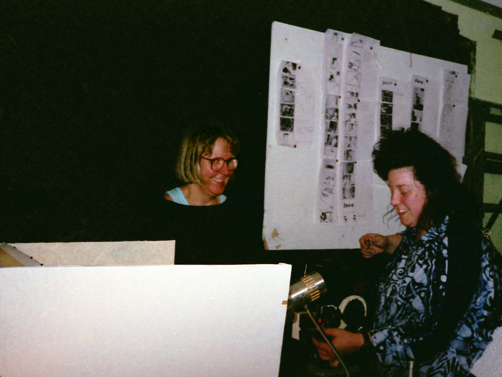 Two women laugh in front of a board of pinned storyboards; one holds a small lamp.