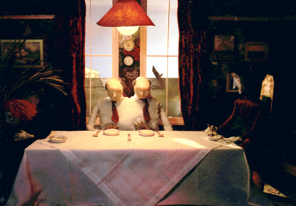 Two identical bald figures with blank faces, in white shirts and red ties, at a laid table.