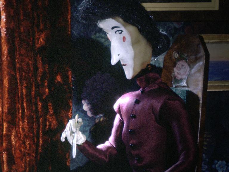 Close view of a black-haired puppet woman with a white face, in maroon, holding a blade.