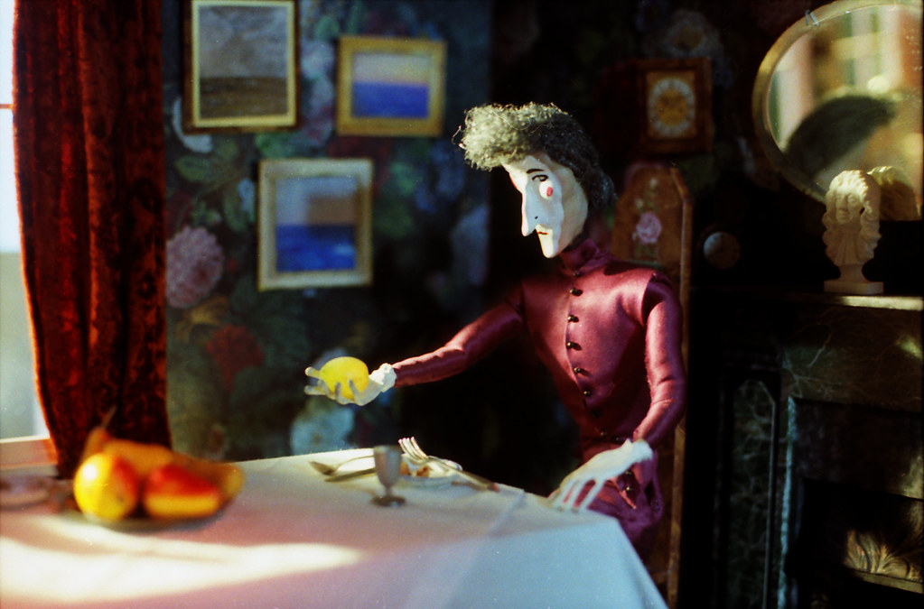A puppet woman in a maroon dress reaches across the table with a yellow fruit on a plate.