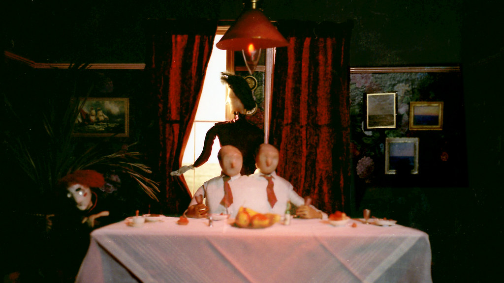 A tall puppet woman in black stands behind two bald figures seated at a dining table.
