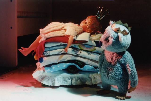 Still from Urpo and Turpo, a doll in a gold crown lying on a stack of folded laundry, Urpo the grey bear standing beside it in a paper crown
